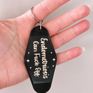 May include: A black motel-style keychain with a diamond shape and ribbed top and bottom edges. The keychain features rose gold metallic text that reads Endometriosis Can Fuck Off, accented by small four-pointed star symbols on both sides.