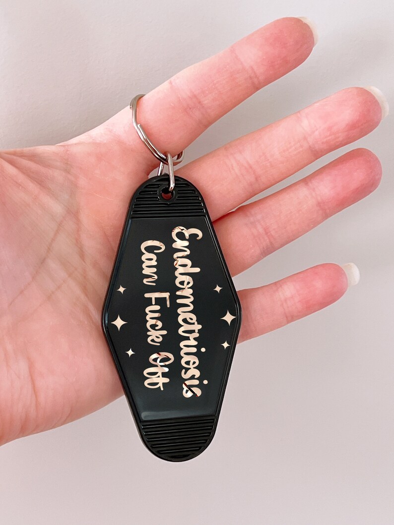 May include: A black motel-style keychain with a diamond shape and ribbed top and bottom edges. The keychain features rose gold metallic text that reads Endometriosis Can Fuck Off, accented by small four-pointed star symbols on both sides.