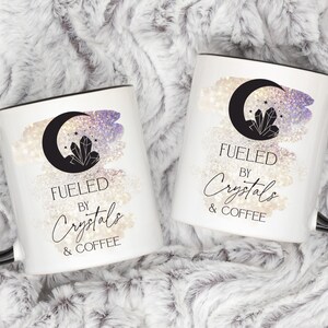 K&ouml;nnte beinhalten: Zwei wei&szlig;e Kaffeetassen mit schwarzen Henkeln und schwarzen R&auml;ndern. Beide Tassen zeigen einen Halbmond mit einem Kristall darin, einen glitzernden Hintergrund und den Text "Fueled by Crystals & Coffee".