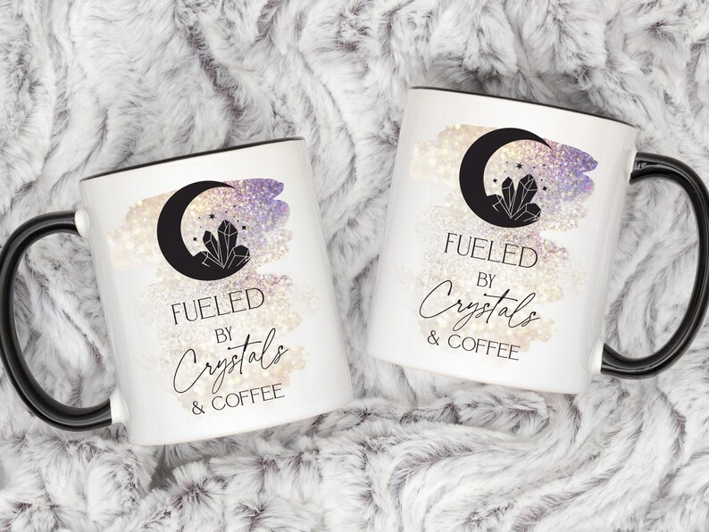 K&ouml;nnte beinhalten: Zwei wei&szlig;e Kaffeetassen mit schwarzen Henkeln und schwarzen R&auml;ndern. Beide Tassen zeigen einen Halbmond mit einem Kristall darin, einen glitzernden Hintergrund und den Text "Fueled by Crystals & Coffee".