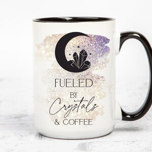 K&ouml;nnte beinhalten: Wei&szlig;e Keramiktasse mit schwarzem Rand und Henkel. Die Tasse zeigt eine Schwarzwei&szlig;illustration eines zunehmenden Mondes mit einem Kristall darin. Der Text "Fueled by Crystals & Coffee" ist in Schwarz unter der Illustration aufgedruckt.