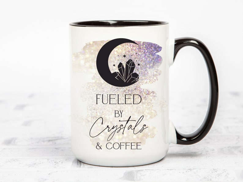 K&ouml;nnte beinhalten: Wei&szlig;e Keramiktasse mit schwarzem Rand und Henkel. Die Tasse zeigt eine Schwarzwei&szlig;illustration eines zunehmenden Mondes mit einem Kristall darin. Der Text "Fueled by Crystals & Coffee" ist in Schwarz unter der Illustration aufgedruckt.