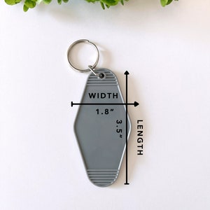 May include: A grey motel-style keychain with a silver metal ring. The keychain features a diamond shape with horizontal ribbed detailing at the top and bottom. Text on the keychain indicates a width of 1.8 inches and a length of 3.5 inches.