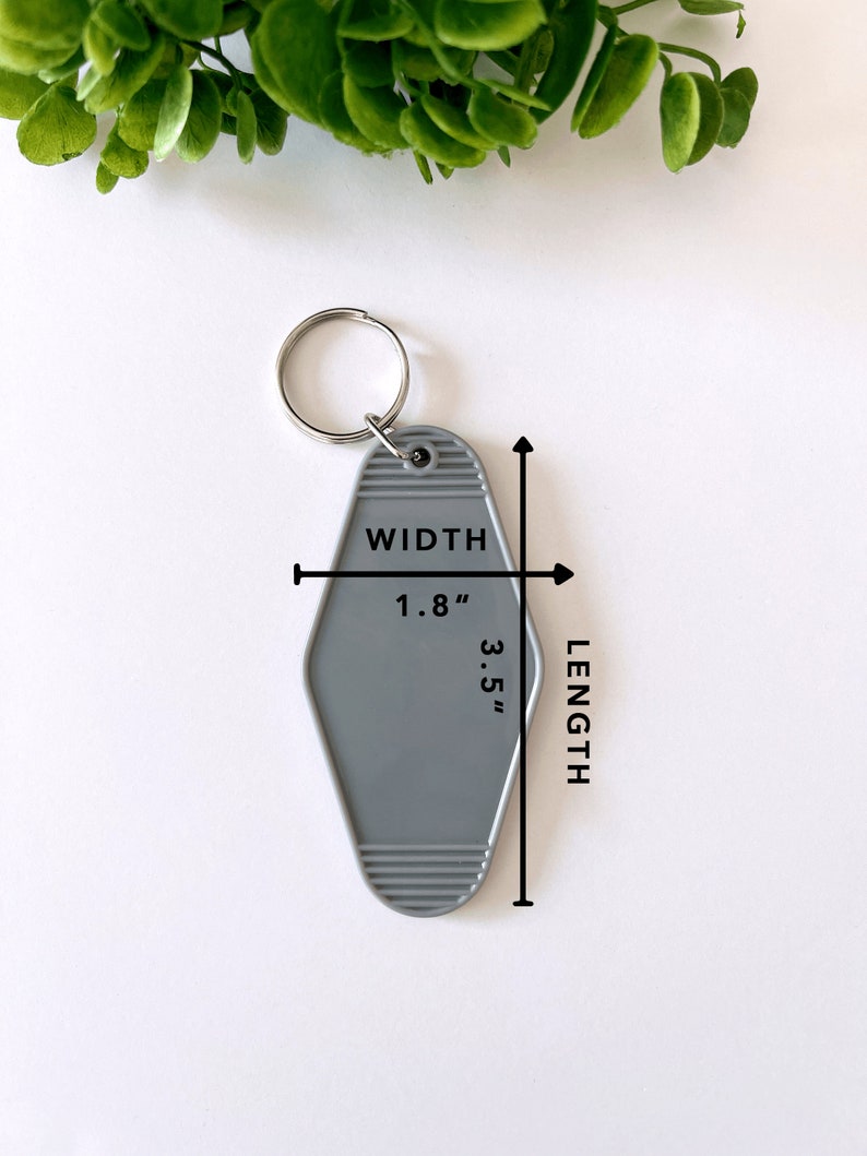 May include: A grey motel-style keychain with a silver metal ring. The keychain features a diamond shape with horizontal ribbed detailing at the top and bottom. Text on the keychain indicates a width of 1.8 inches and a length of 3.5 inches.