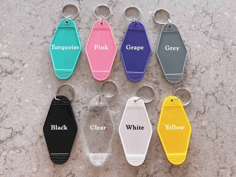 May include: Eight motel-style keychains arranged in two rows. The top row features turquoise, pink, grape, and grey colors. The bottom row includes black, clear, white, and yellow. Each keychain has a metal ring and horizontal ribbed details.