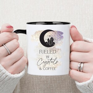 K&ouml;nnte beinhalten: Wei&szlig;e Keramik-Kaffeetasse mit schwarzem Rand und Henkel. Die Tasse zeigt eine Schwarzwei&szlig;illustration eines zunehmenden Mondes mit Kristallen und Sternen. Der Text "Fueled by Crystals & Coffee" ist in schwarzer Kursivschrift gedruckt.