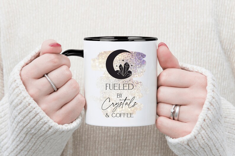 K&ouml;nnte beinhalten: Wei&szlig;e Keramik-Kaffeetasse mit schwarzem Rand und Henkel. Die Tasse zeigt eine Schwarzwei&szlig;illustration eines zunehmenden Mondes mit Kristallen und Sternen. Der Text "Fueled by Crystals & Coffee" ist in schwarzer Kursivschrift gedruckt.