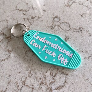 May include: A teal motel-style keychain with a silver metal ring. The keychain features the text "Endometriosis Can Fuck Off" in light pink lettering, accented by small four-pointed star shapes, with ribbed detailing on the left and right ends.