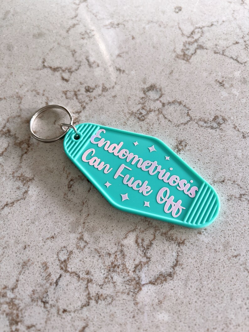 May include: A teal motel-style keychain with a silver metal ring. The keychain features the text "Endometriosis Can Fuck Off" in light pink lettering, accented by small four-pointed star shapes, with ribbed detailing on the left and right ends.