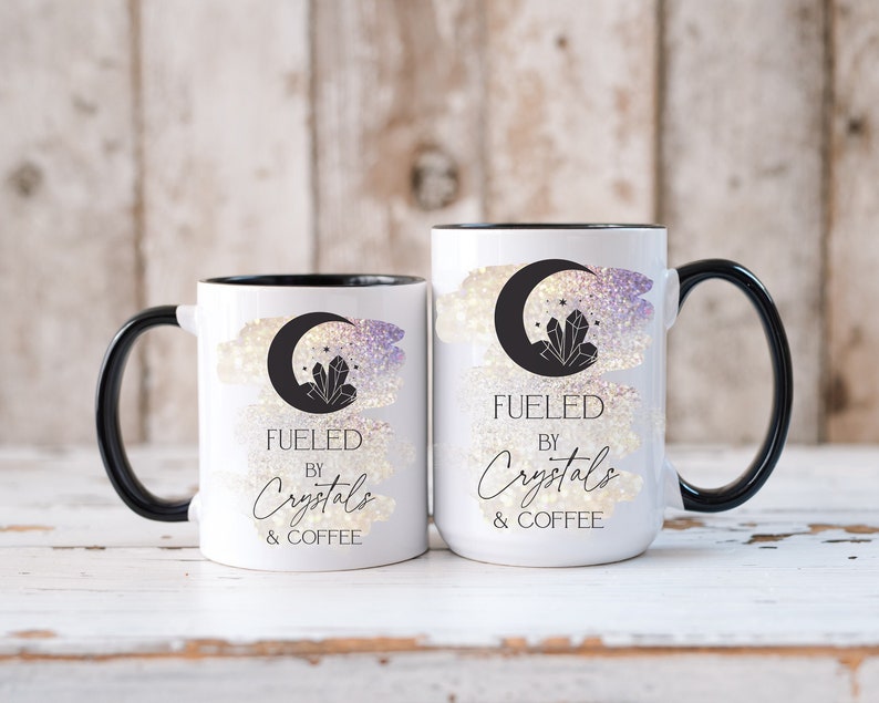 K&ouml;nnte beinhalten: Zwei wei&szlig;e Keramikbecher mit schwarzen R&auml;ndern und Griffen. Die Becher haben ein Aquarell-Design mit einer Mondsichel und Kristallen. Der Text auf den Bechern lautet "Fueled by Crystals & Coffee".