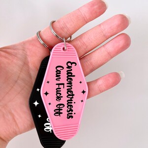 May include: A pink motel-style keychain with black text that reads Endometriosis Can Fuck Off and small star icons, attached to a silver metal ring. A second black keychain with white text is partially visible behind the pink one.