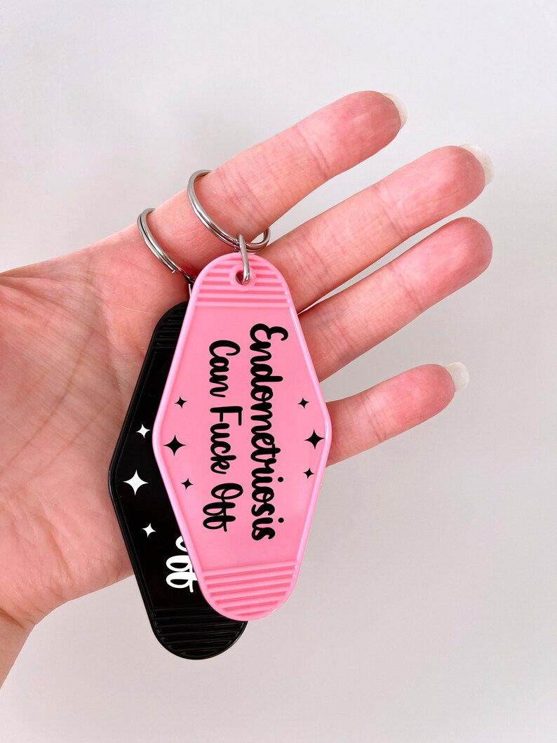 May include: A pink motel-style keychain with black text that reads Endometriosis Can Fuck Off and small star icons, attached to a silver metal ring. A second black keychain with white text is partially visible behind the pink one.