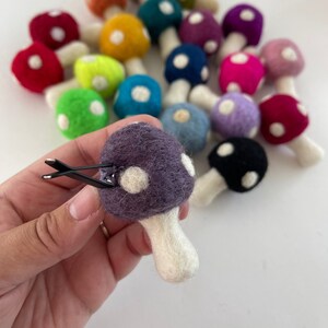 May include: A collection of felt mushroom hair clips in various colors, including red, green, blue, purple, and black. Each mushroom has white polka dots and a white stem. The mushrooms are arranged in a circle, with a purple mushroom in the center.