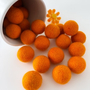 May include: A white ceramic cup spills out orange felt balls onto a white surface. The felt balls are soft and round, and they are scattered around the cup.