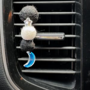 May include: A car air vent clip featuring a black and white felt ball design, a silver star, a white felt ball, and a blue crescent moon charm. The clip is designed to attach to a car's air vent.