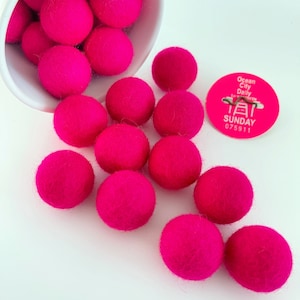 May include: A white bowl filled with bright pink felt balls, spilling out onto a white surface. The felt balls are approximately 1 inch in diameter.
