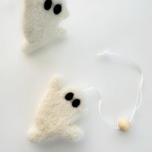 May include: Two white felt ghost ornaments with black eyes. One ghost has a white string attached to a small wooden bead. The ghosts are handmade and have a soft, textured appearance. Perfect for Halloween decorations.