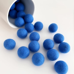 May include: A collection of bright blue felt balls spilling from a white ceramic container. The balls are scattered on a white surface, creating a playful and visually appealing arrangement. The felt balls are a vibrant shade of blue.