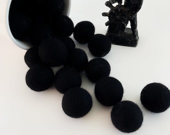 May include: A white ceramic cup spills out black felt balls onto a white surface. A small black metal figurine of a person turning a wheel is to the right of the felt balls.