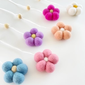 May include: A collection of colorful felt flower ornaments in shades of pink, purple, blue, peach, and white. Each flower has a contrasting center and is strung on a white cord with wooden beads. The flowers are arranged on a white surface.
