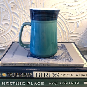 May include: A ceramic pitcher with a handle, in shades of blue and teal, sits atop a stack of books. The top rim is dark blue, transitioning to lighter shades. The books include "The Old Man and the Sea" by Hemingway.