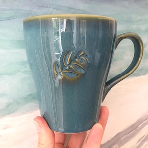 May include: A teal ceramic mug with a leaf design. The mug has a glossy finish with a light green rim and handle. The leaf design is embossed and has a gold color.