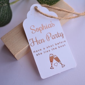 May include: A white rectangular paper tag with rounded scalloped edges and a jute twine loop. The tag features rose gold foil text that reads Sophia's Hen Party, Have a shot before she ties the knot, and an illustration of two clinking glasses.