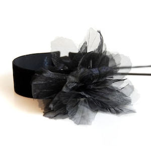Könnte beinhalten: Ein schwarzer Samt-Choker mit einem großen, geschichteten, durchsichtigen schwarzen Blumenakzent. Die Blume besteht aus zartem, fließendem Stoff, und der Choker liegt vor einem weißen Hintergrund.