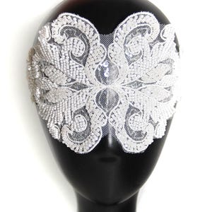 Op de afbeelding: Een witte, met kralen bezette oogmasker met een ingewikkeld bloemenontwerp. Het masker is gemaakt van delicate kant en is versierd met kleine, glinsterende kralen. Het masker wordt getoond op een zwarte mannequinhoofd tegen een witte achtergrond.