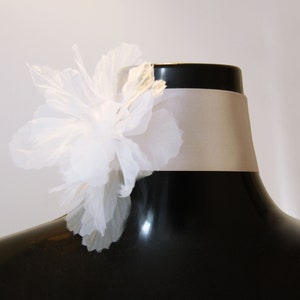 Könnte beinhalten: Ein weißes, mehrschichtiges Stoffblumen-Accessoire verziert ein Satinband-Choker. Der Choker hat eine hellbeige Farbe und wird auf einer schwarzen Schaufensterpuppe präsentiert. Die Blume hat zarte, transparente Blütenblätter.