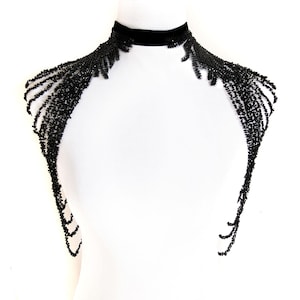 Könnte beinhalten: Ein schwarzes, mit Perlen besetztes Schulterstück mit einem Samt-Choker. Die Perlenstränge fallen über Schultern und Arme. Das Stück wird auf einer weißen Schaufensterpuppe präsentiert, was einen auffälligen Kontrast erzeugt.
