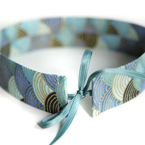 Könnte beinhalten: Ein Stoff-Halsband mit einem sich wiederholenden Muster aus überlappenden Halbkreisen in Blau-, Hellblau- und Goldtönen. Das Band wird vorne mit einem dünnen, salbeigrünen Satinband zu einer kleinen Schleife gebunden.