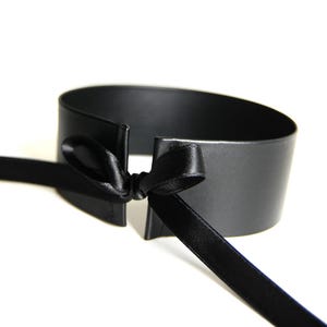 Puede incluir: Un brazalete negro con una cinta de satén atada con un lazo. El brazalete está hecho de un material mate y tiene un diseño simple y elegante. La cinta añade un toque de sofisticación.