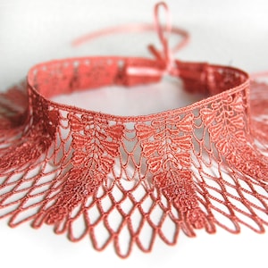 Könnte beinhalten: Ein korallenfarbenes Spitzen-Halsband mit einem komplizierten, sich wiederholenden blattartigen Muster am oberen Rand und einem zarten, durchbrochenen Rautennetz-Design, das nach unten verläuft. Das Stück enthält ein dünnes, passendes Band zum Binden.