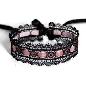 May include: Black lace choker necklace with a pink ribbon detail. The choker has a scalloped lace design and a black bow closure. A delicate accessory for adding a touch of elegance.