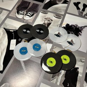 Puede incluir: Un conjunto de seis pendientes de vinilo negro y blanco en forma de disco. Dos pendientes presentan un diseño de espiral negro y blanco, dos pendientes presentan un diseño de espiral negro y blanco con un centro azul y dos pendientes presentan un disco de vinilo negro con un centro amarillo y la palabra "beat" impresa en negro.