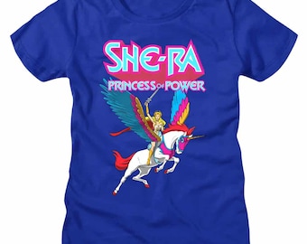 May include: Blue t-shirt featuring the text "SHE-RA PRINCESS OF POWER" in pink and teal. The graphic shows She-Ra riding a winged unicorn with a sword. The unicorn is white with a pink mane and tail.