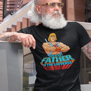 May include: Black t-shirt featuring a cartoon image of a muscular man with a sword and the text "GREATEST FATHER IN THE UNIVERSE" in blue, red, and orange. The shirt is worn by a person with a white beard and sunglasses.