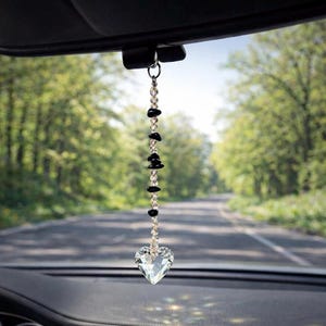 May include: A car charm with a clear crystal heart pendant. The charm is suspended from a silver-colored ring and features a strand of clear beads and black stones. The background shows a road and trees.