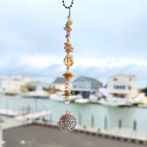 May include: A hanging ornament featuring a silver-toned metal sand dollar charm suspended from a vertical strand of assorted beige and cream-colored stone chips, clear faceted crystal beads, and a single larger amber-colored glass bead.