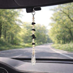 May include: A car charm featuring a clear crystal pendant with a faceted design, suspended from a chain of small, clear beads and black stones. The charm hangs from a silver ring attached to the car's interior, with a blurred road and trees in the background.