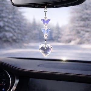 May include: A car charm featuring a clear, faceted heart pendant suspended below two butterfly charms with purple and blue accents. The charm hangs from the rearview mirror, with a snowy outdoor scene visible in the background.