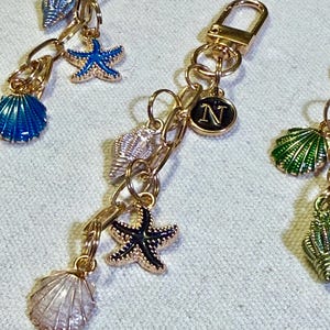 May include: Gold-toned metal keychains featuring dangling charms of seashells and starfish in various colors, including blue, green, and white. Each keychain includes a circular charm with the letter N engraved in gold on a black background.