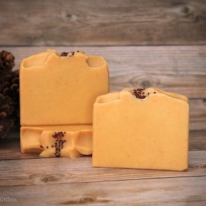 May include: Two bars of handmade soap, one larger than the other, are displayed on a wooden surface. Both bars are a light orange color and have a decorative design on top. The soap is decorated with small brown seeds.