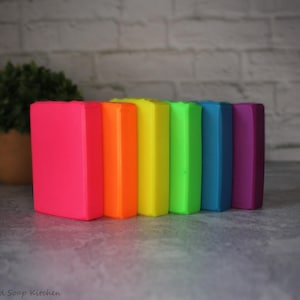 May include: A row of seven vibrantly colored rectangular soaps in a rainbow spectrum: pink, orange, yellow, green, blue, and purple. The soaps are arranged on a gray surface with a brick wall background and a potted plant.
