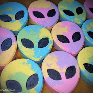 May include: A collection of colorful, alien-shaped bath bombs. Each bomb features a pastel color scheme with a black oval eye design. The bath bombs are in shades of pink, yellow, blue, and green, with a speckled, glittery finish.