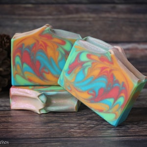May include: Three bars of handmade soap with a swirl pattern in shades of orange, green, blue, and red. The soap is on a wooden surface.