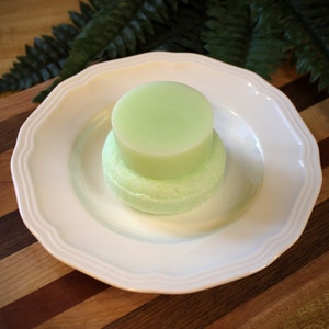 May include: A green and white solid shampoo bar on a white plate. The bar is round and has a smooth surface.