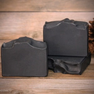 May include: Three bars of black charcoal soap stacked on top of each other on a wooden surface. The soap is handmade and has a rough, textured surface.