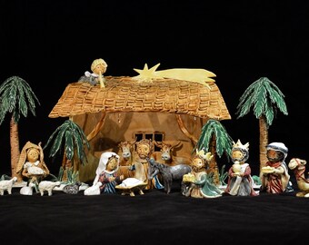 Puede incluir: Un belén de cerámica con un establo de techo de paja marrón, palmeras, una estrella y figuras de María, José, el niño Jesús, los tres reyes magos, pastores, animales y ovejas.